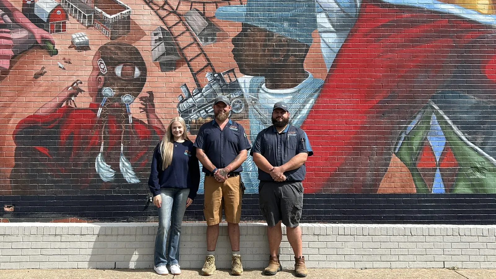 The Good Times Plumbing crew in their navy work shirts, standing in front of the railroad mural in downtown Conway