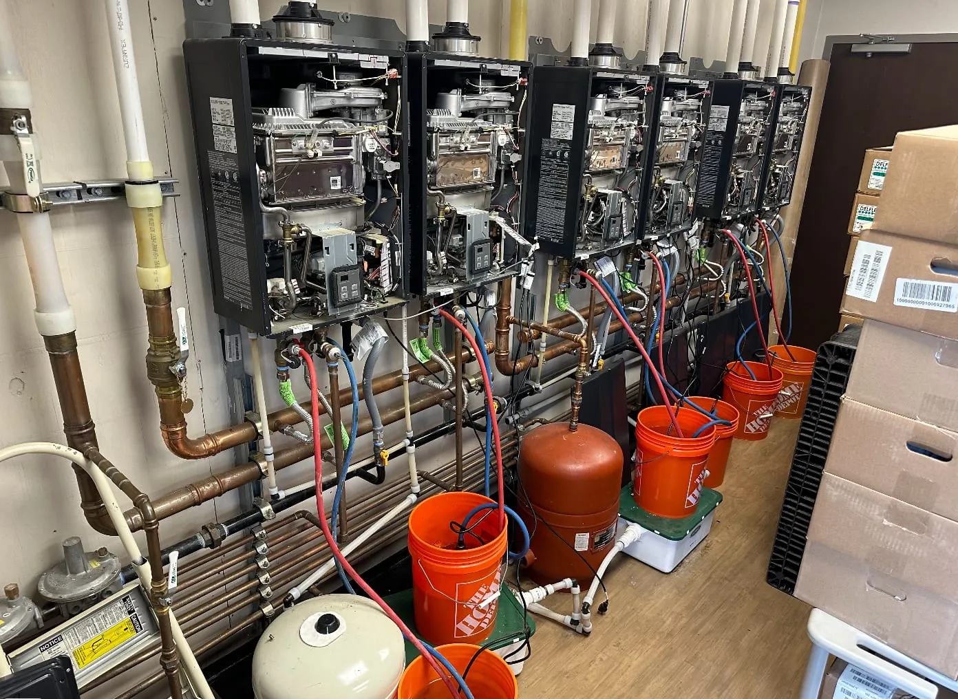 A row of tankless water heaters mid-flush, with descaling pumps and lines connected by Good Times Plumbing