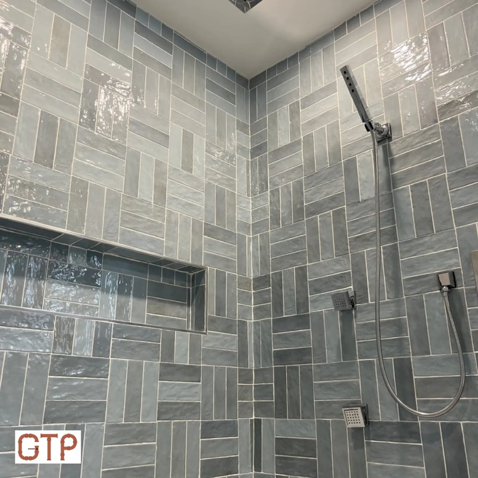 A finished custom tile shower by Good Times Plumbing with herringbone-set tile, a rain head, and body sprays